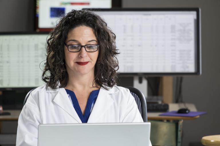 Woman working on a laptop with computer monitors in the background.