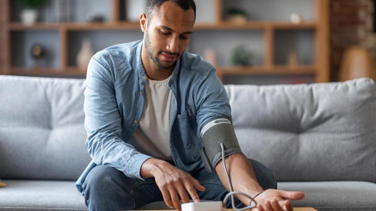Photo of young African-American man sitting on a couch and checking blood pressure with upper arm cuff and monitor.